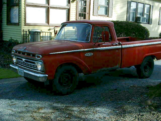 Faded red 1966-style Ford F-series pickup parked in driveway beside yellow house, front quarter view