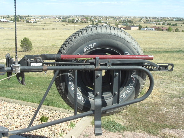Swing-away spare tire carrier with Goodyear Wrangler tire and red jack, rural background