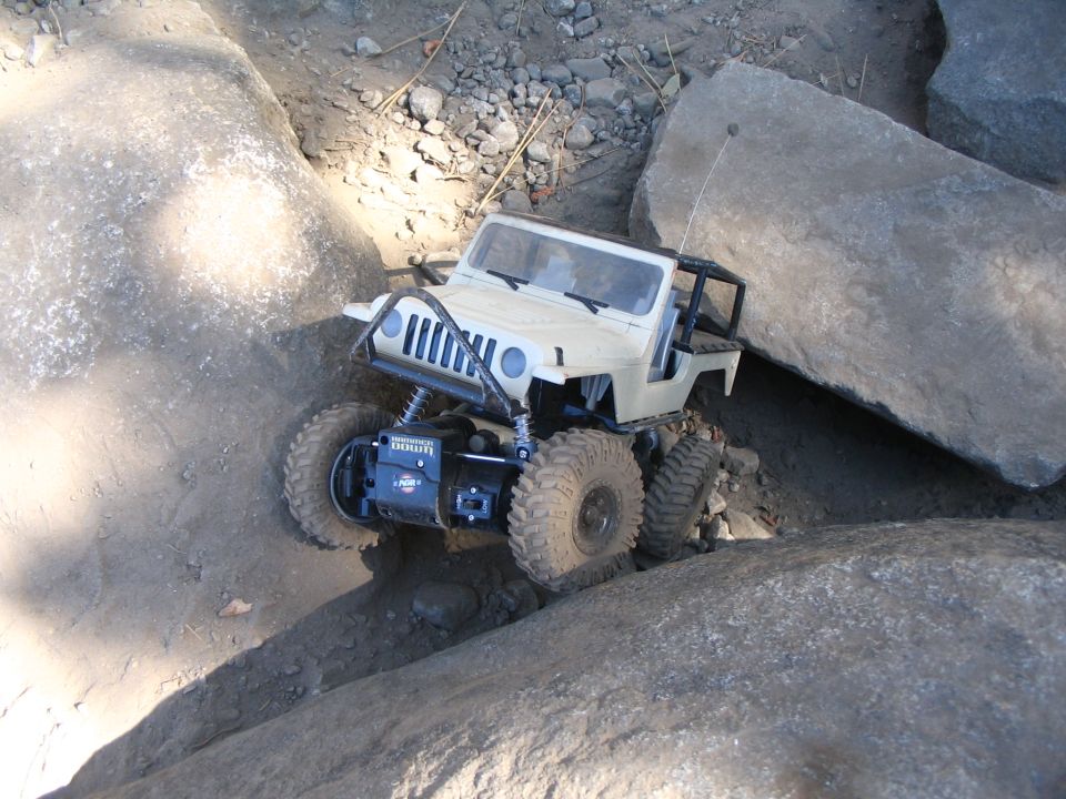 Tan RC crawler nose-down between rocks, exposed motor and winch, muddy tires