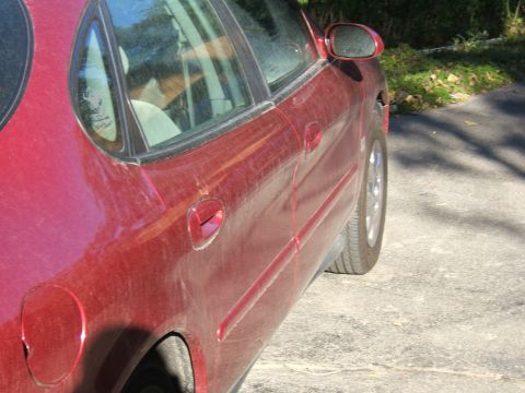 1998 Ford Taurus - The side of a red sedan is shown, covered in dust, with a partial view of the interior and a side mirror.