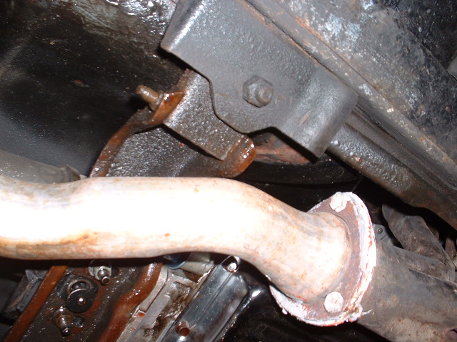 Exhaust pipe flange with white sealant and rusty bolts, attached to a black metal bracket