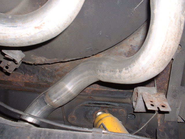 Underside view of a vehicle's exhaust pipes and rusty frame components. A yellow shock absorber is visible