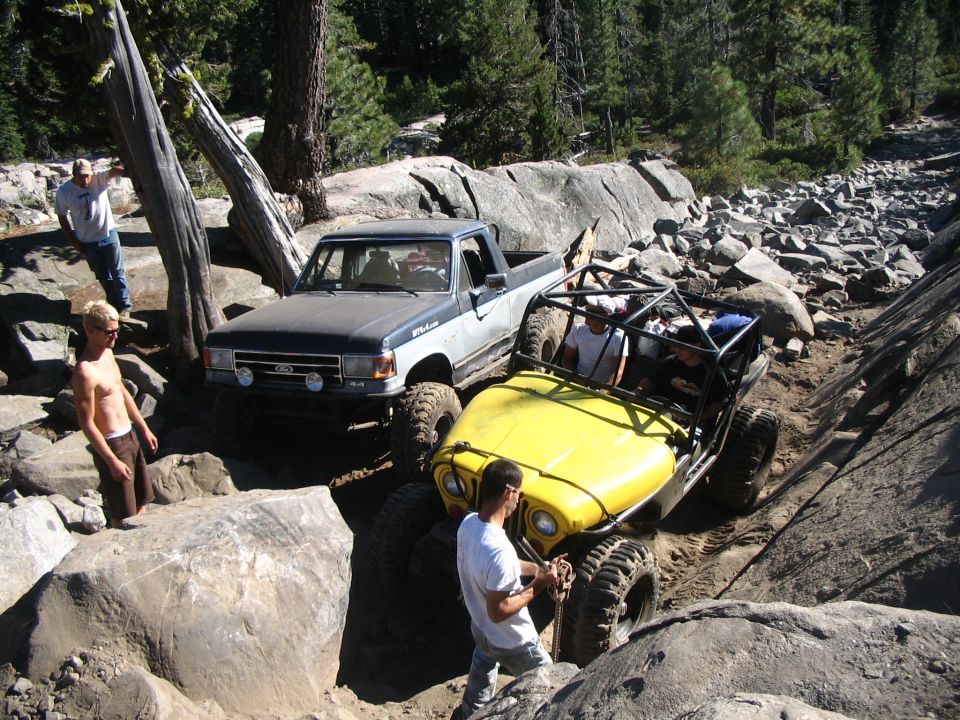 Yellow tube buggy and blue Ford pickup crawling over large boulders on a forested rock trail