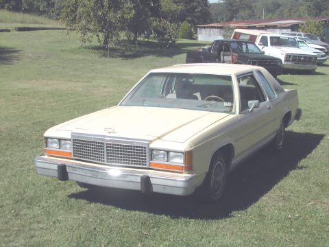 1985 Ford LTD Crown Victoria - A tan Ford sedan is parked on a grassy area with several other vehicles and buildings visible in the background.