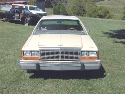 1985 Ford LTD Crown Victoria - A cream-colored sedan is parked in a grassy yard with two trucks in the background.