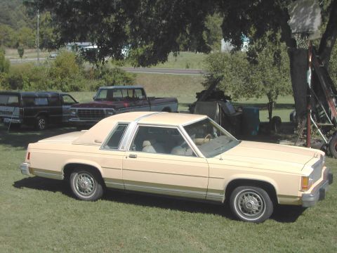 1985 Ford LTD Crown Victoria - A tan Ford sedan is parked in a yard with other vehicles and trees in the background.