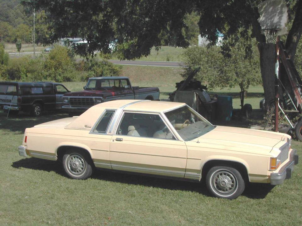 Tan Ford sedan parked on grass with two pickup trucks behind it
