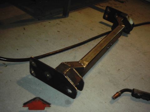 1978 Ford Bronco - A fabricated metal bracket sits on a concrete floor next to a welding torch tip and a red arrow marker.