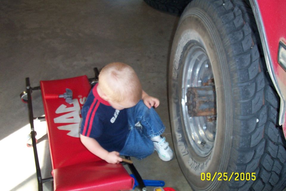 Toddler in jeans and a striped shirt sits on a red creeper next to a large truck tire