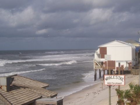 1995 Ford Bronco - A beach scene with a restaurant on stilts under a stormy sky, with waves crashing on the shore.