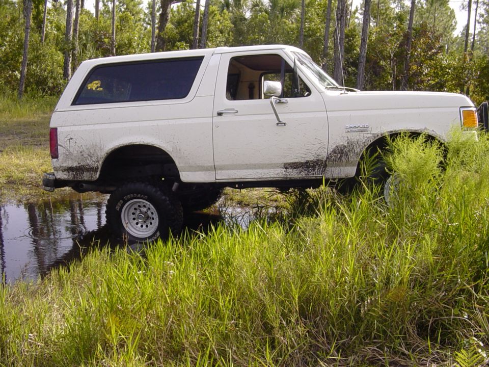 1989 Ford Bronco driving through a muddy puddle in a grassy, wooded area