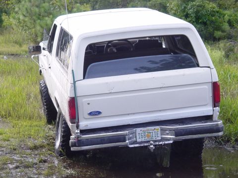 1989 Ford Bronco - A white Ford Bronco is partially submerged in mud and water, viewed from the rear.