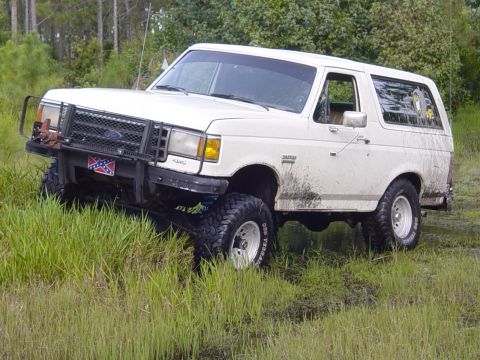 1989 Ford Bronco - White Ford Bronco with brush guard and lifted suspension driving through tall grass.