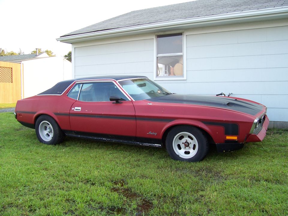 Red coupe with black hood and roof parked on grass next to a house. Looks like a 1973 Mustang
