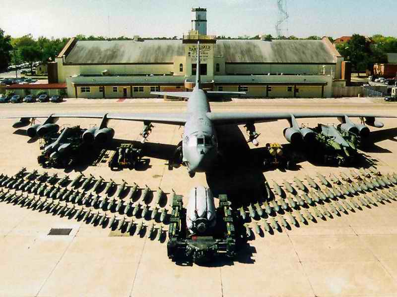 Bomber aircraft on tarmac with rows of bombs and a building in the background