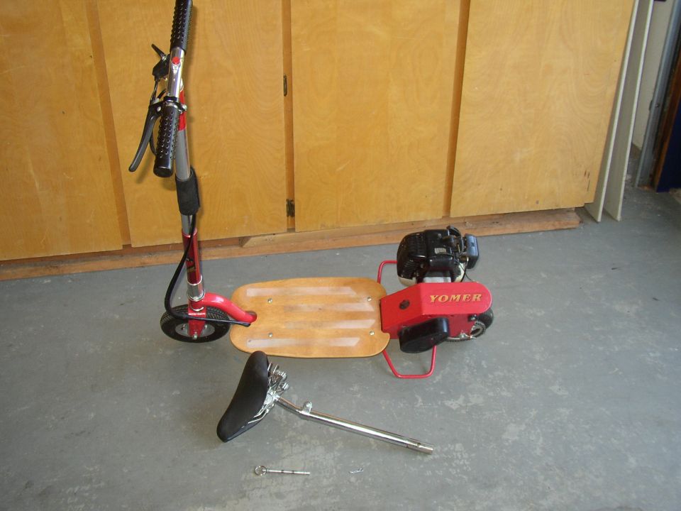 Red motorized scooter with a wooden deck, detached seat, and a Yomer engine. The scooter is on a concrete floor in front of wooden cabinets