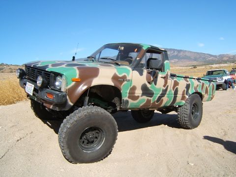 1988 Ford Bronco - A pickup truck with a camouflage paint scheme is parked on a dirt road with other vehicles in the background.