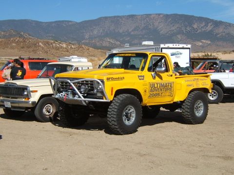 1988 Ford Bronco - Modified yellow pickup truck with off-road tires and graphics, parked with other vehicles in a desert landscape.