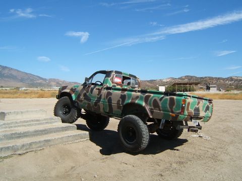 1988 Ford Bronco - A lifted pickup truck with a camouflage paint scheme is shown on a dirt lot with concrete steps.