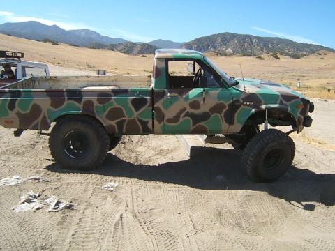 1988 Ford Bronco - Camouflaged pickup truck with "4WD" marking sits on sandy ground under a clear sky.