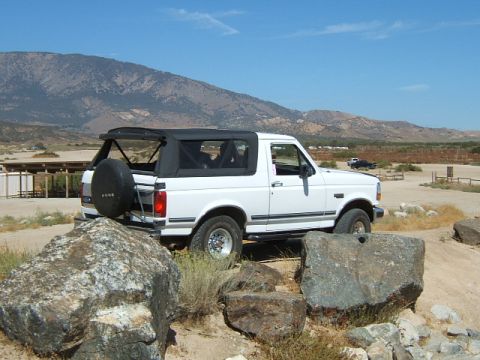 1988 Ford Bronco - A white Ford Bronco with a black soft top is parked on a dirt lot with mountains in the background.