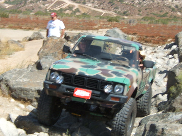 Camouflage pickup truck with large tires navigating rocky terrain. A person stands in the background