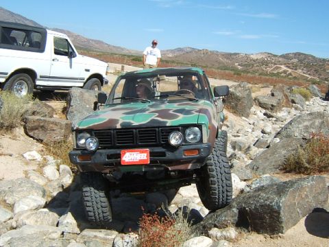 1988 Ford Bronco - A camouflaged pickup truck is shown off-roading over rocks, with a white Bronco visible behind it.