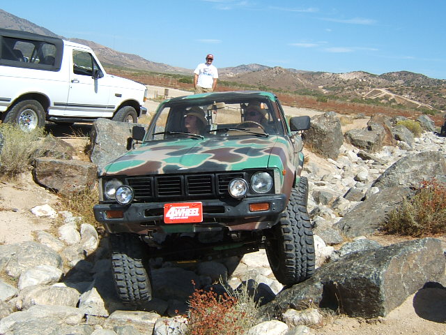 Camouflaged pickup truck navigating rocks, with a white Bronco in the background