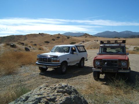 1988 Ford Bronco - Two Ford Broncos, one from the first generation and a later model, parked on a dusty trail with arid hills in the background.