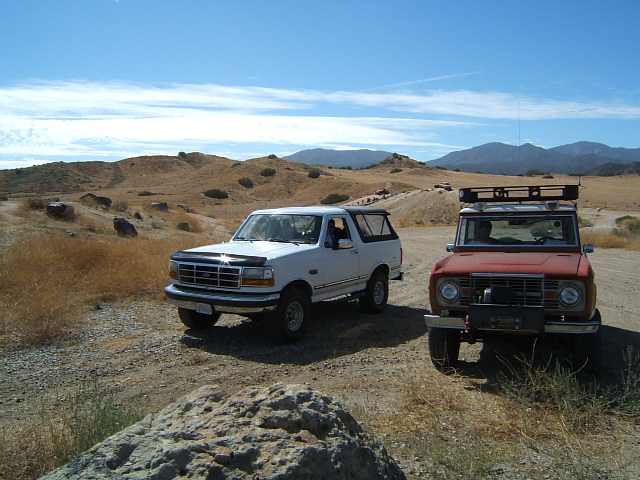Red first-generation Ford Bronco with roof rack and white fourth-generation Ford Bronco on dirt road