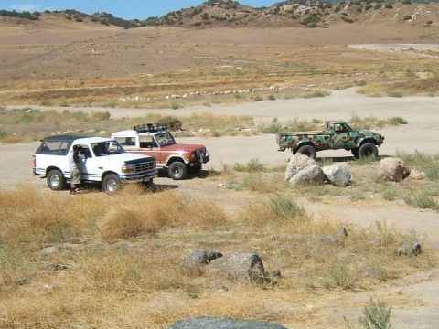 1988 Ford Bronco - Three off-road vehicles are parked in a dry, open area with hills in the background under a clear blue sky.