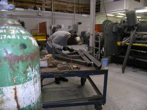 2001 Jeep Wrangler - A welder works on a metal frame in a workshop, with a large green tank and industrial machinery visible in the background.