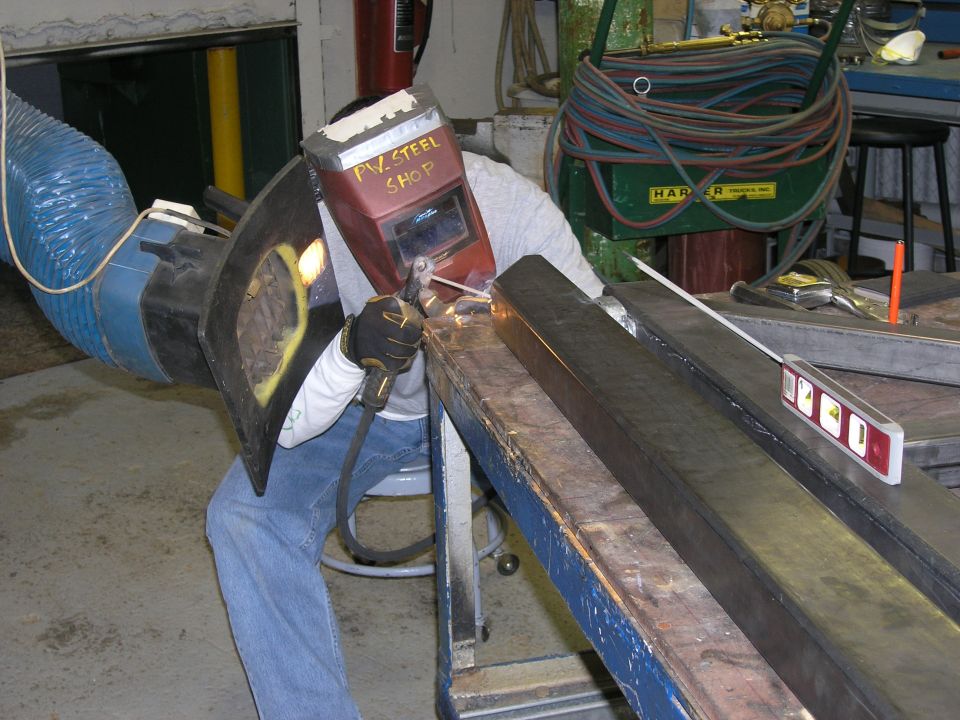 Welder in mask and gloves working on metal beams with welding torch. Blue exhaust hose visible