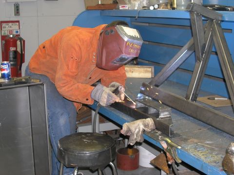 2001 Jeep Wrangler - A welder is actively working on a metal frame, sparks flying as they weld. The welder wears protective gear, including a mask with 'PV Stee…
