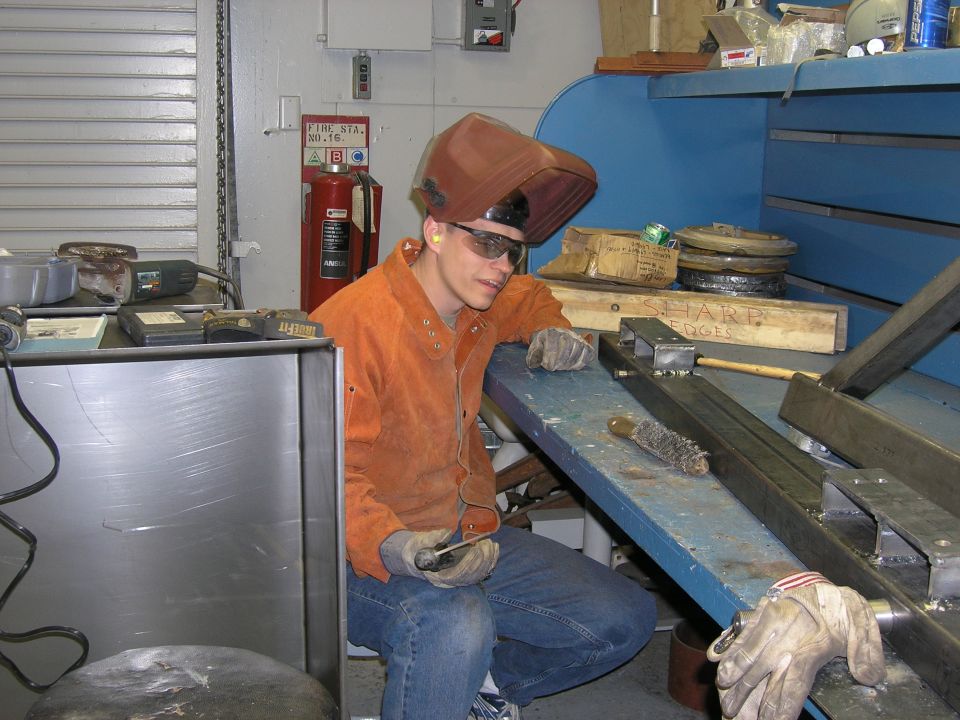 Welder wearing a mask and safety glasses, leaning on a workbench with metal parts