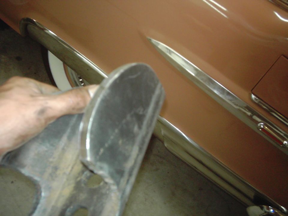 Hand holding a metal part near the side of a tan car with chrome trim. A white wall tire is visible