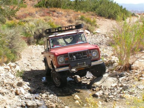 1988 Ford Bronco - This red Ford Bronco navigates a challenging rocky stream, equipped with a roof rack for adventure.