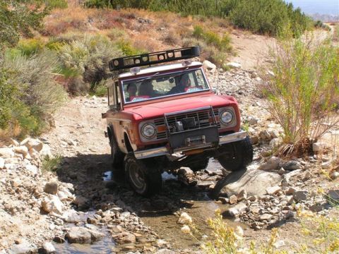 1988 Ford Bronco - First-generation Ford Bronco tackles an off-road trail with rocky terrain and a stream crossing.
