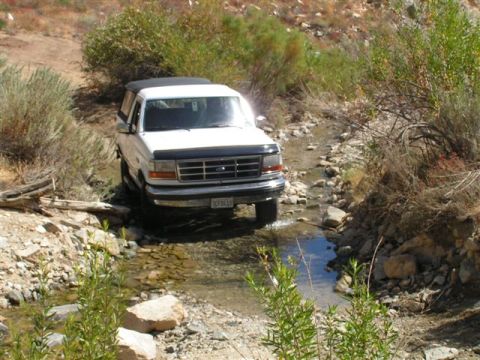 1988 Ford Bronco - A white Ford Bronco navigates a dry creek bed, its front end angled towards the viewer.