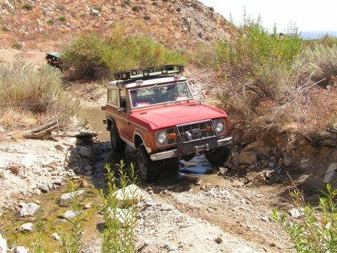 1988 Ford Bronco - A red first-generation Ford Bronco tackles an off-road trail, splashing through a shallow stream with another vehicle in the distance.