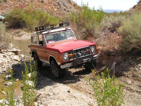 1988 Ford Bronco - Red Ford Bronco navigates a rocky off-road trail with brush and hills surrounding it.