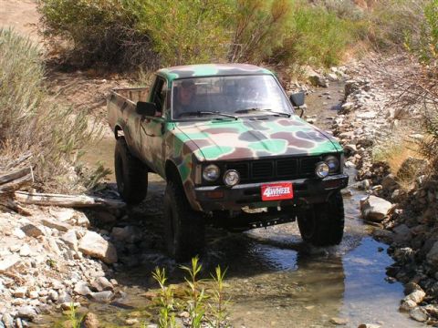 1988 Ford Bronco - A camouflage pickup truck navigates a rocky stream bed in a dry, brushy environment.