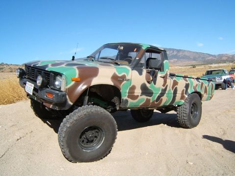 1973 Ford Bronco - Modified pickup truck with camouflage paint and off-road tires sits on a dusty track under a clear blue sky.