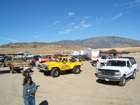 1973 Ford Bronco - A group of off-road vehicles are gathered in a dusty lot under a clear blue sky, with people milling around.