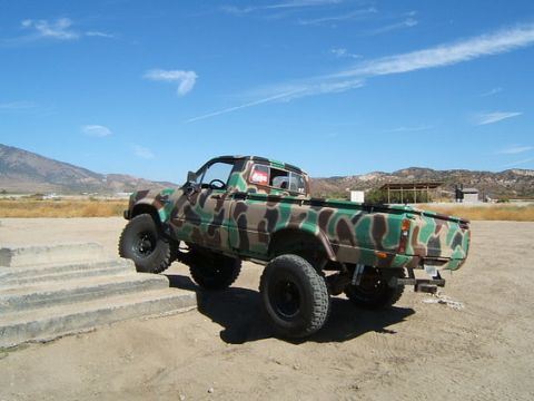 1973 Ford Bronco - A camouflaged pickup truck with off-road modifications is positioned on a dirt surface near concrete steps under a clear blue sky.