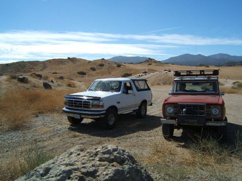 1973 Ford Bronco - Two Ford Broncos are parked on a dusty trail in a dry, hilly landscape under a blue sky with scattered clouds.