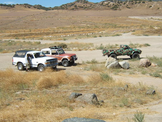 White 1995 Ford Bronco, orange Ford Bronco, and camouflage pickup truck parked in a dry, hilly landscape