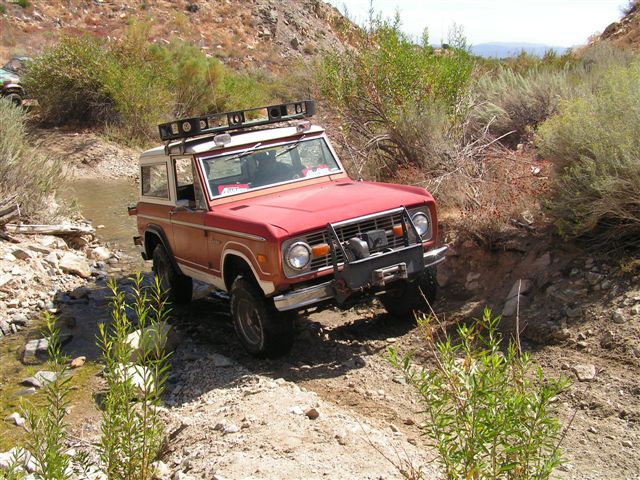 Red 1973 Ford Bronco navigating a rocky stream bed with a roof rack
