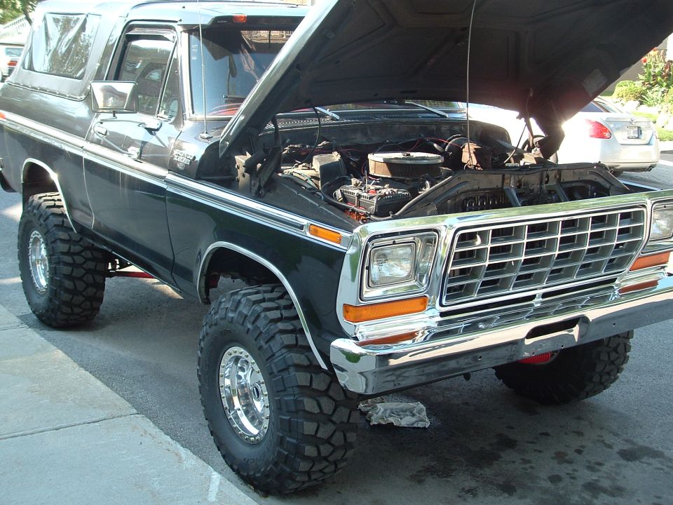 Dark green lifted Ford Bronco with hood up, chrome grille, large off-road tires, in driveway