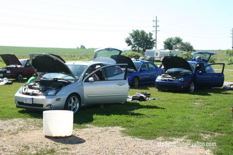 2002 Ford Focus - Several cars, including a silver Ford Focus and two blue Subaru WRXs, are parked on a grassy field with their hoods open.